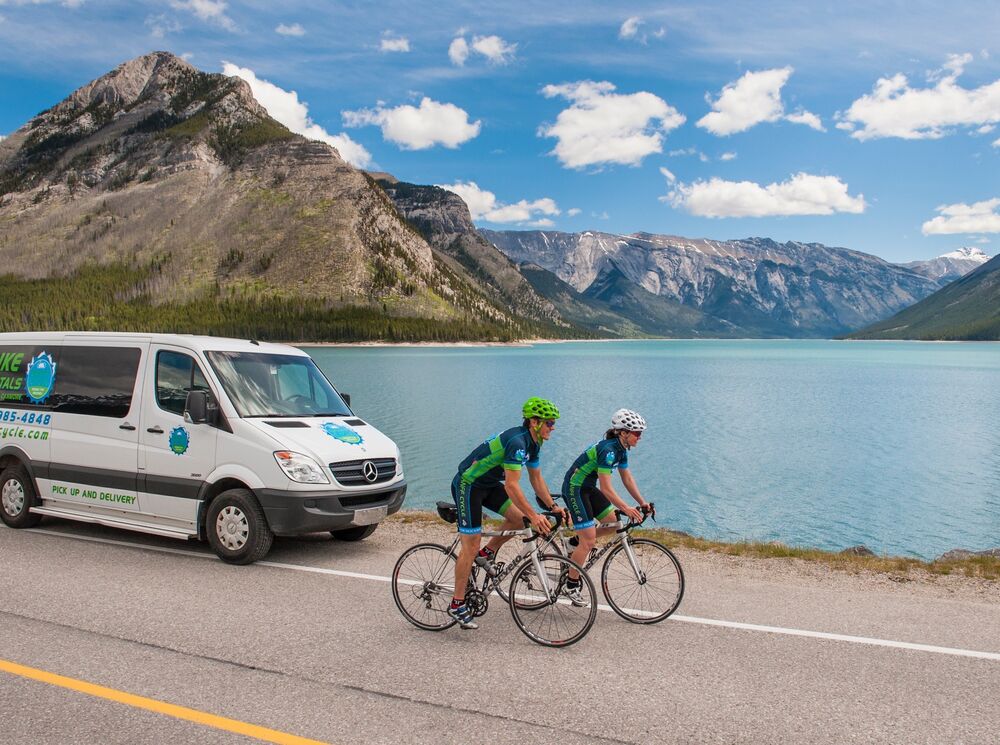 Two cyclists and a support vehicle ride along the Minnewanka Loop in Banff National Park on a bluebird day with a mountain behind them.