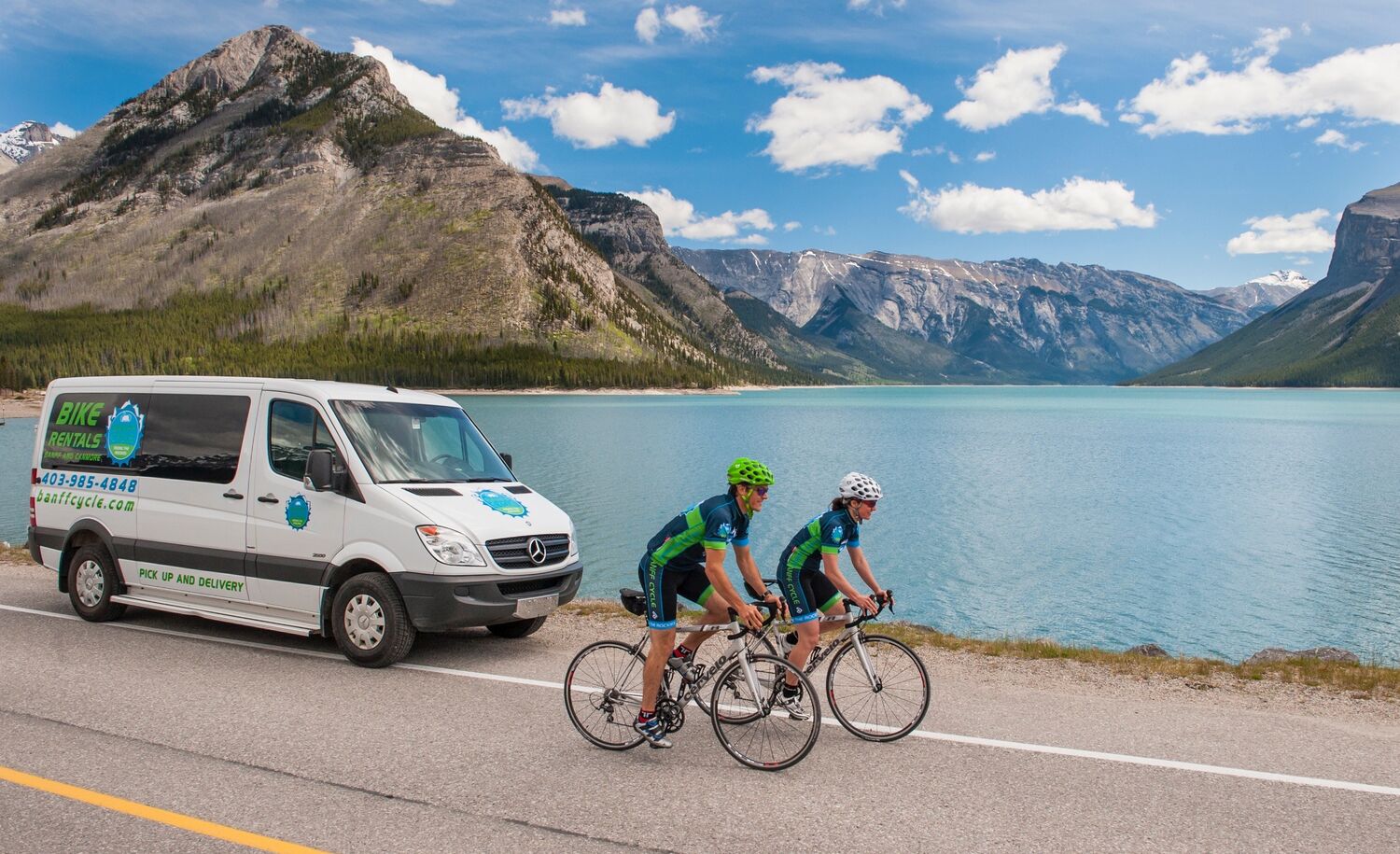 Two cyclists and a support vehicle ride along the Minnewanka Loop in Banff National Park on a bluebird day with a mountain behind them.