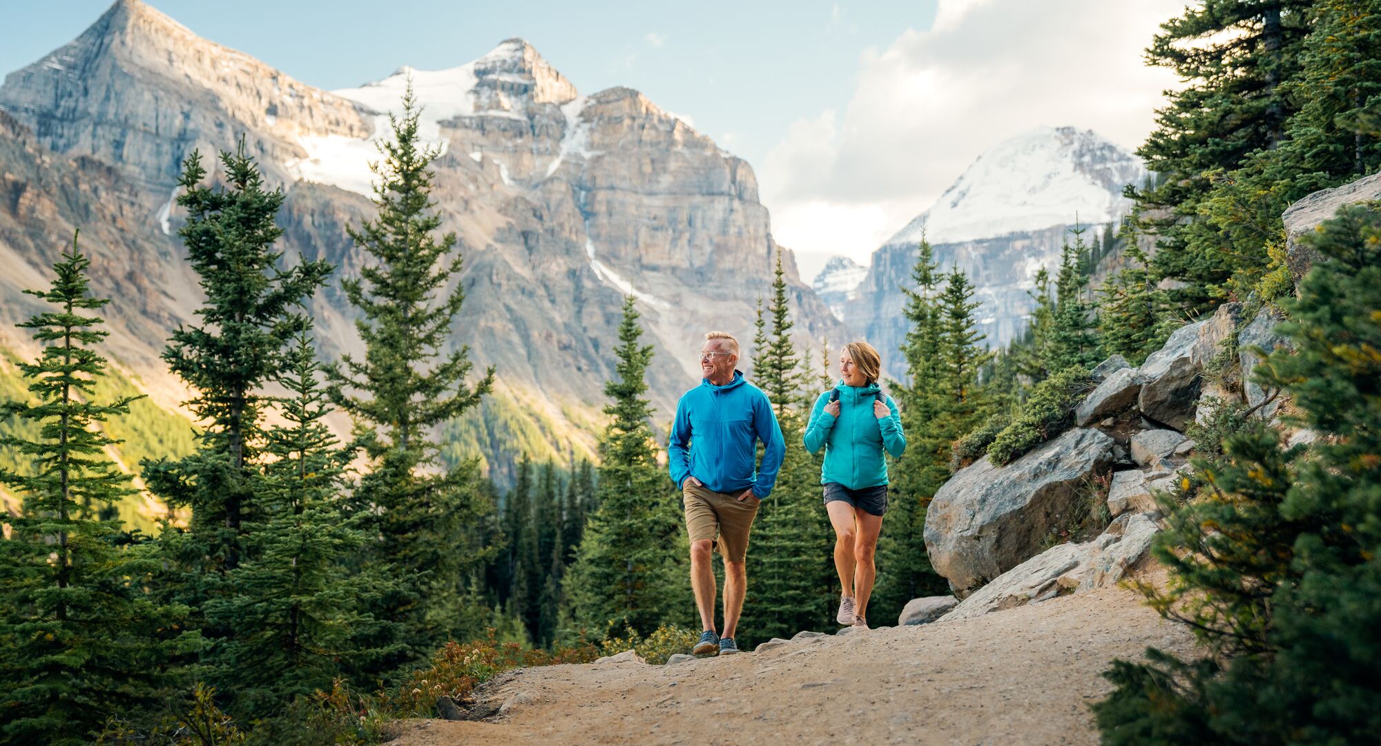 Two people hiking in Banff National Park on a trail at Lake Louise in the summer with mountains behind them.
