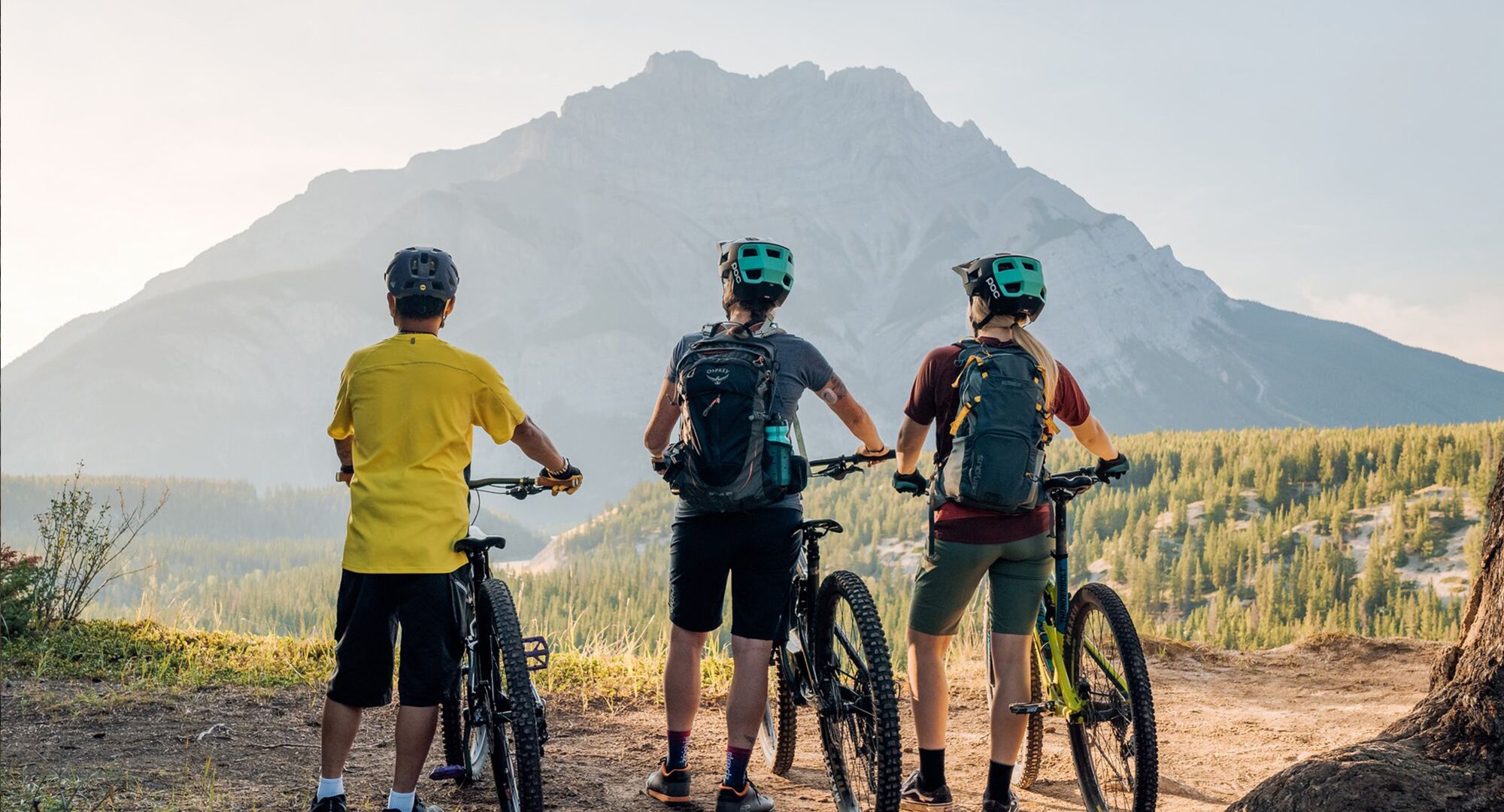 Three people stand at the top of a mountain biking route in Banff National Park looking out at Mount Rundle in the summer.