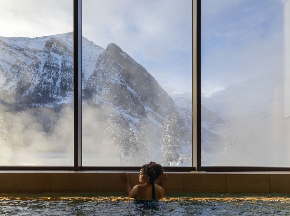A woman in a hot tub looks out a window at a snowy mountain at BASIN Glacier Spa in Lake Louise in Banff National Park.