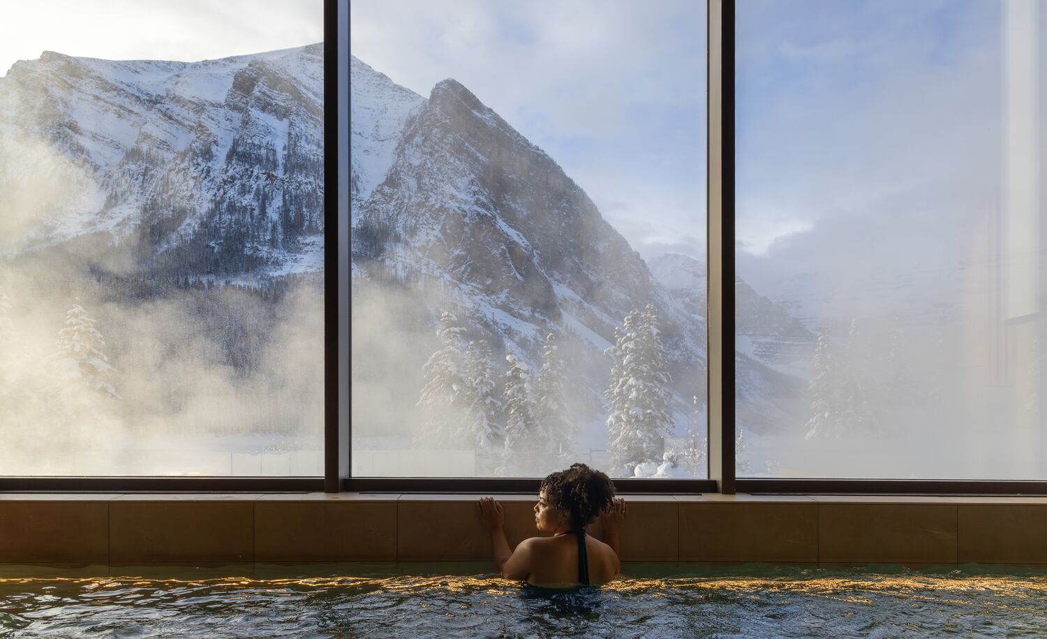 A woman in a hot tub looks out a window at a snowy mountain at BASIN Glacier Spa in Lake Louise in Banff National Park.