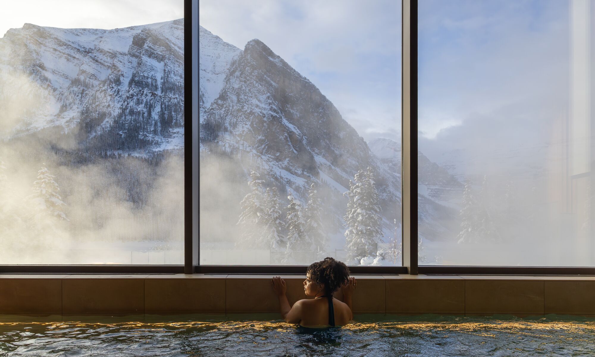 A woman in a hot tub looks out a window at a snowy mountain at BASIN Glacier Spa in Lake Louise in Banff National Park.