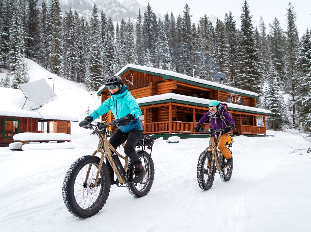 Two cyclists ride e-fat bikes away from Sundance Lodge in the winter while biking in Banff National Park.