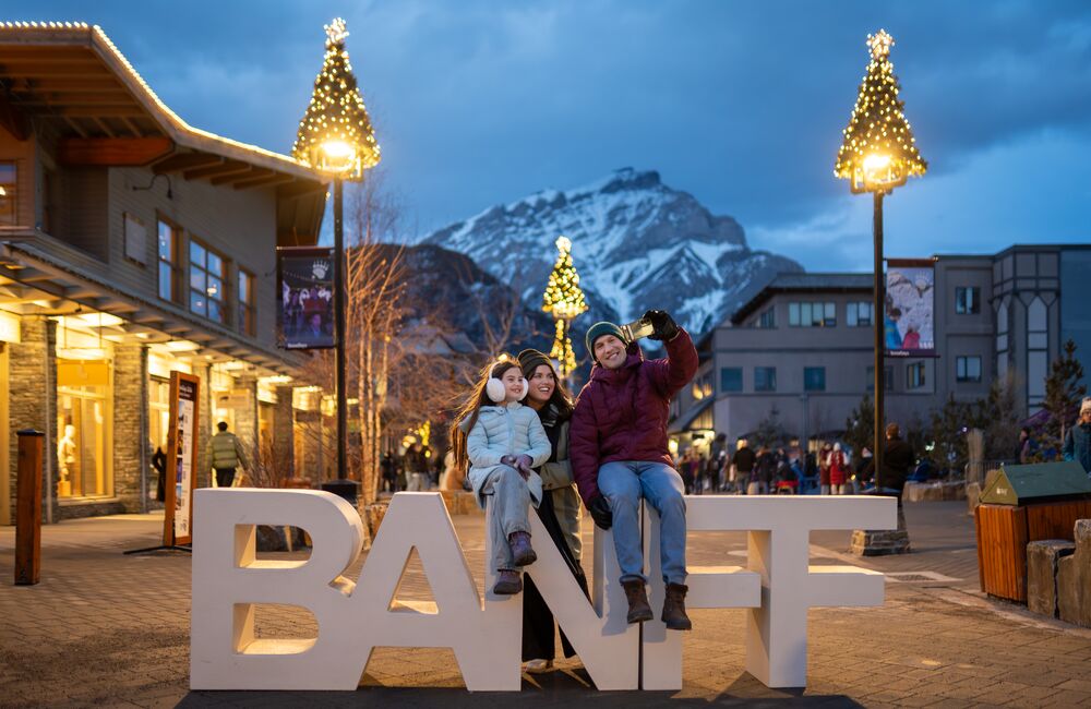 Family posing at the iconic Banff sign in downtown Banff during the Art of Ice Carving Competition event at SnowDays Festival, one of the top winter events in Banff National Park, surrounded by stunning ice sculptures and snowy mountain scenery