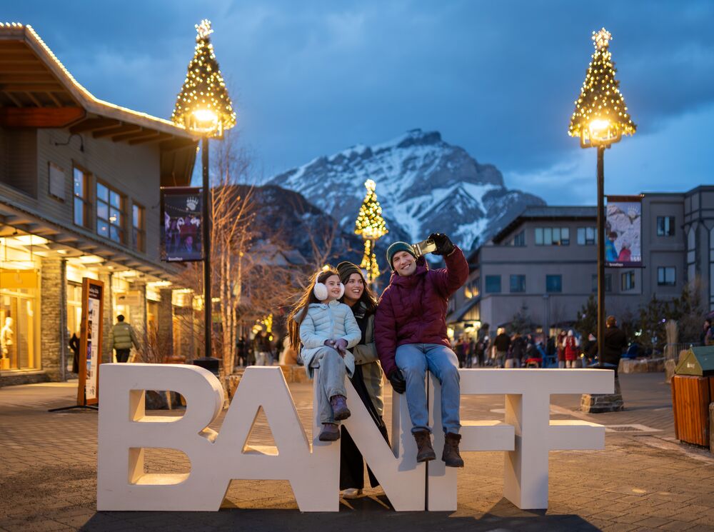 Family posing at the iconic Banff sign in downtown Banff during the Art of Ice Carving Competition event at SnowDays Festival, one of the top winter events in Banff National Park, surrounded by stunning ice sculptures and snowy mountain scenery