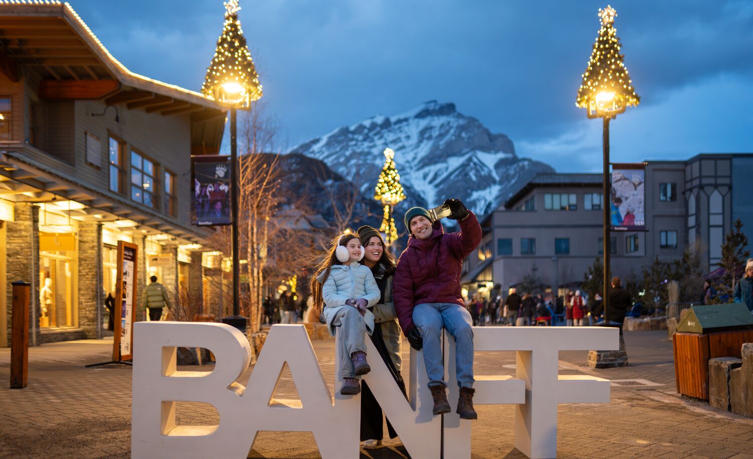 Family posing at the iconic Banff sign in downtown Banff during the Art of Ice Carving Competition event at SnowDays Festival, one of the top winter events in Banff National Park, surrounded by stunning ice sculptures and snowy mountain scenery