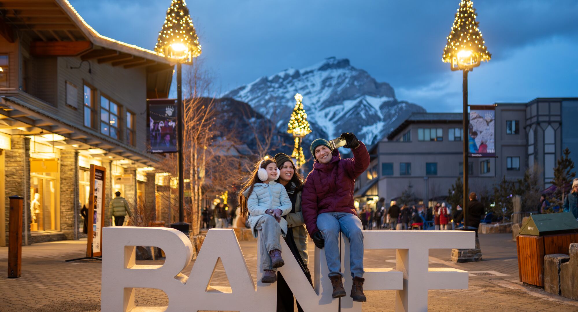 Family posing at the iconic Banff sign in downtown Banff during the Art of Ice Carving Competition event at SnowDays Festival, one of the top winter events in Banff National Park, surrounded by stunning ice sculptures and snowy mountain scenery