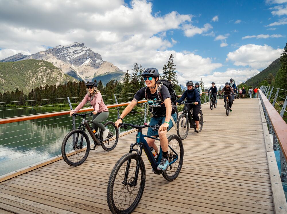 A group of cyclists ride on a pedestrian bridge in Banff on a sunny summer day.