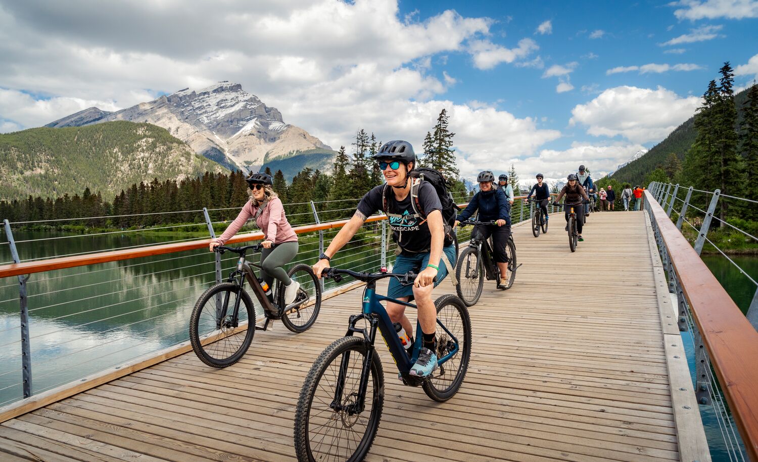 A group of cyclists ride on a pedestrian bridge in Banff on a sunny summer day.