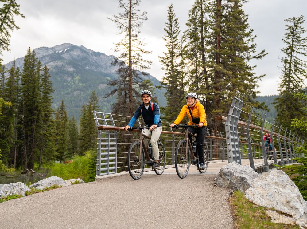 Two cyclists ride across a bridge in the Banff Townsite on a summer day while biking in Banff National Park.