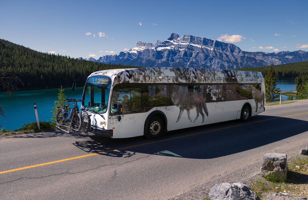A Roam Transit bus drives the Minnewanka Loop in Banff National Park on a blue bird day in front of Two JAck Lake.