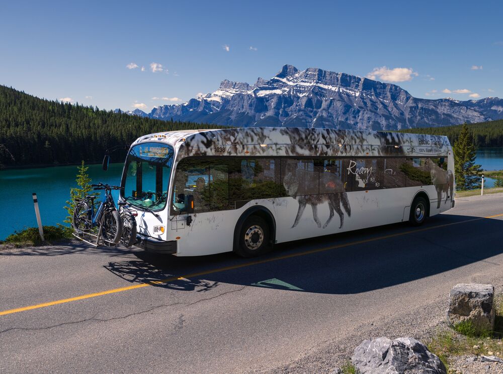 A Roam Transit bus drives the Minnewanka Loop in Banff National Park on a blue bird day in front of Two JAck Lake.