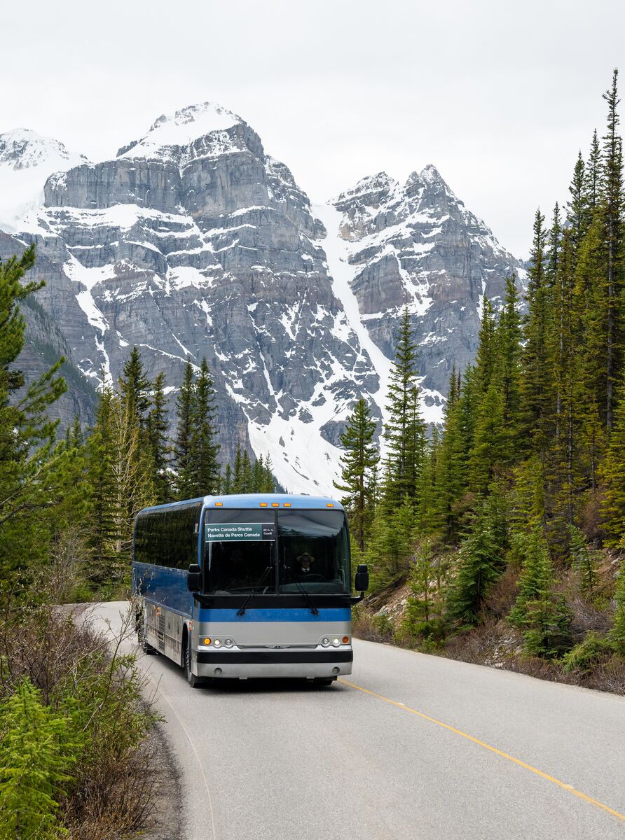 A shuttle bus goes down Moraine Lake Road with mountains behind it in the summer.