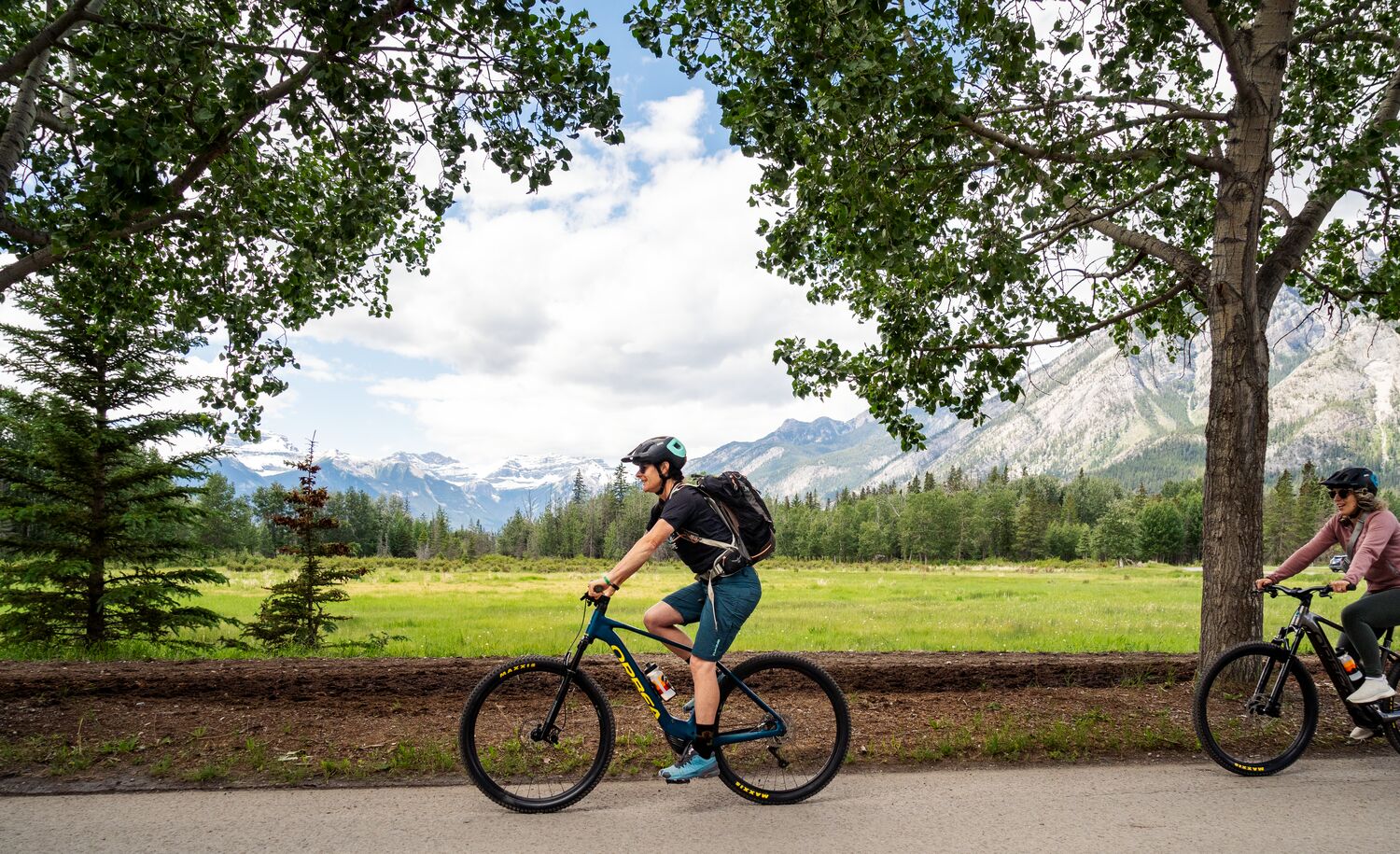 Two people bike along a road in Banff National Park, heading towards the Sundance Trail on a bike tour.