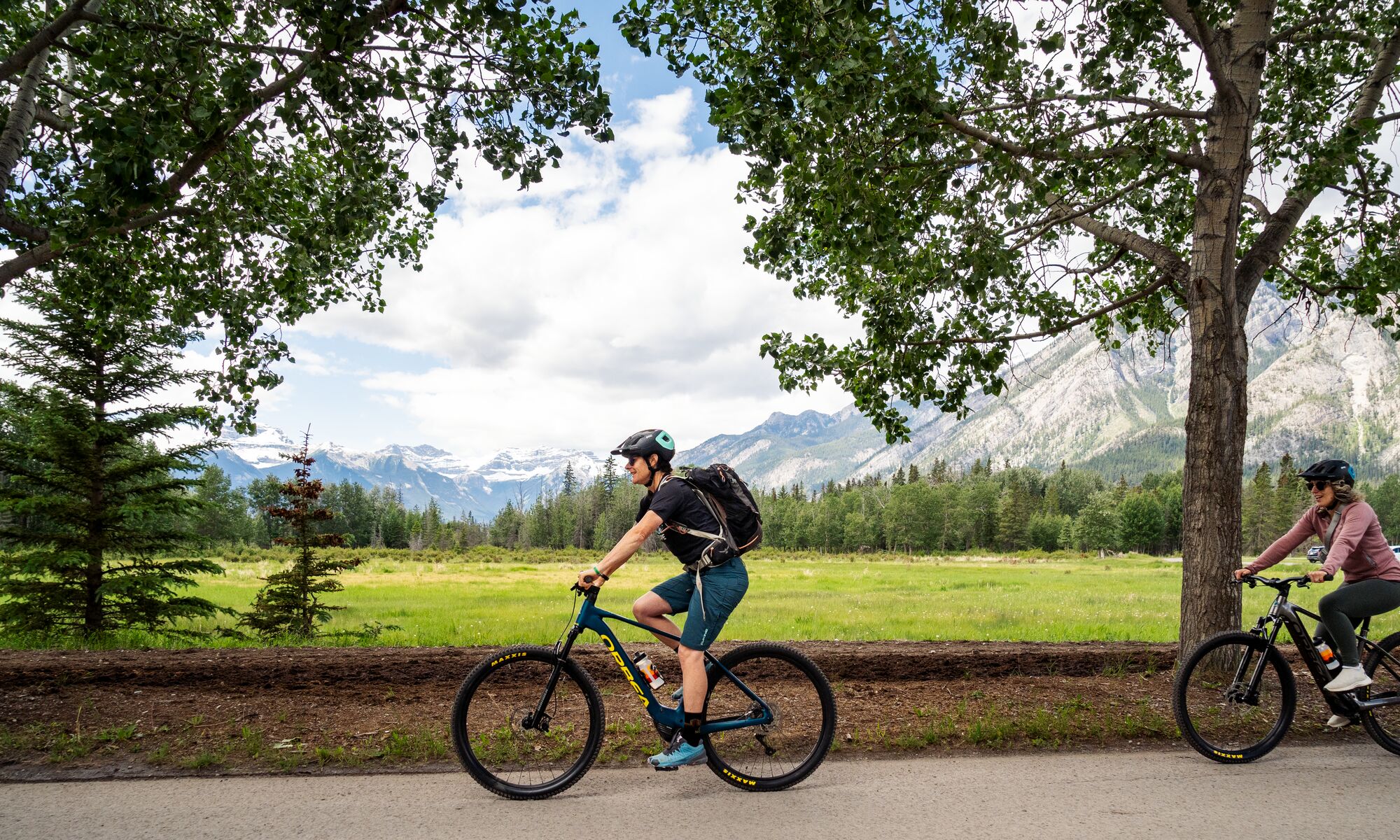 Two people bike along a road in Banff National Park, heading towards the Sundance Trail on a bike tour.