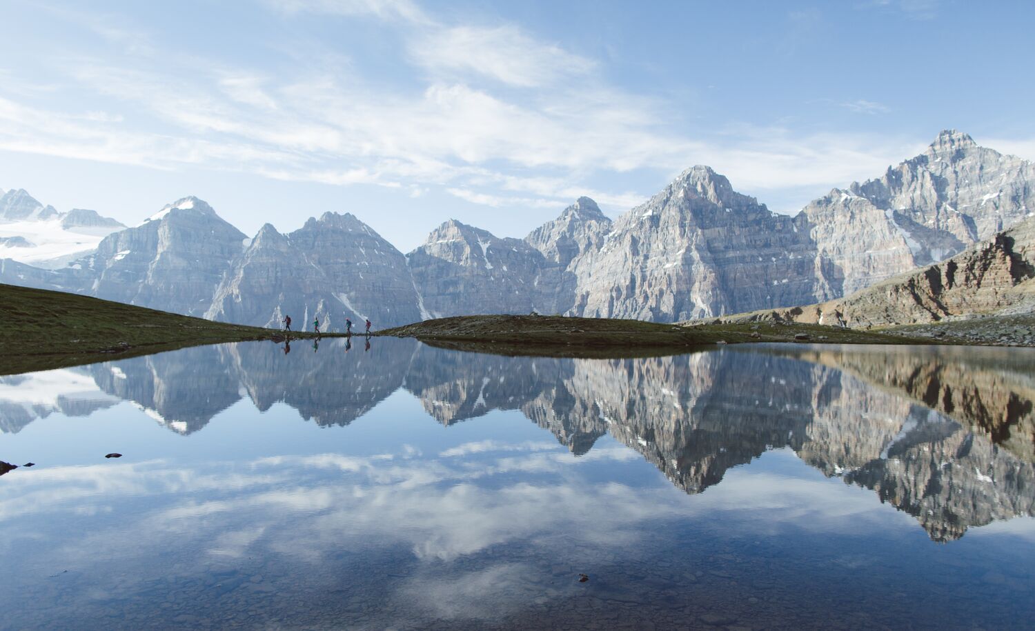 Four people reflected in a lake in Larch Valley with the mountains of the Valley of the Ten Peaks behind them while taking a hike at Moraine Lake in Banff National Park.