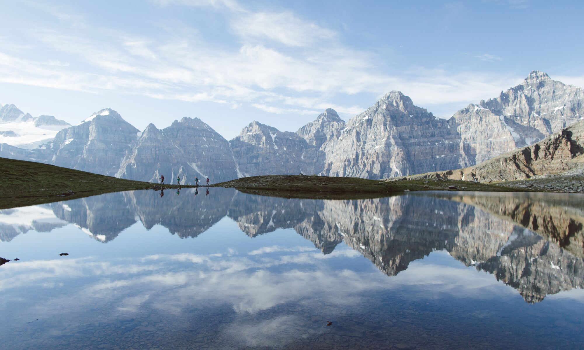 Four people reflected in a lake in Larch Valley with the mountains of the Valley of the Ten Peaks behind them while taking a hike at Moraine Lake in Banff National Park.