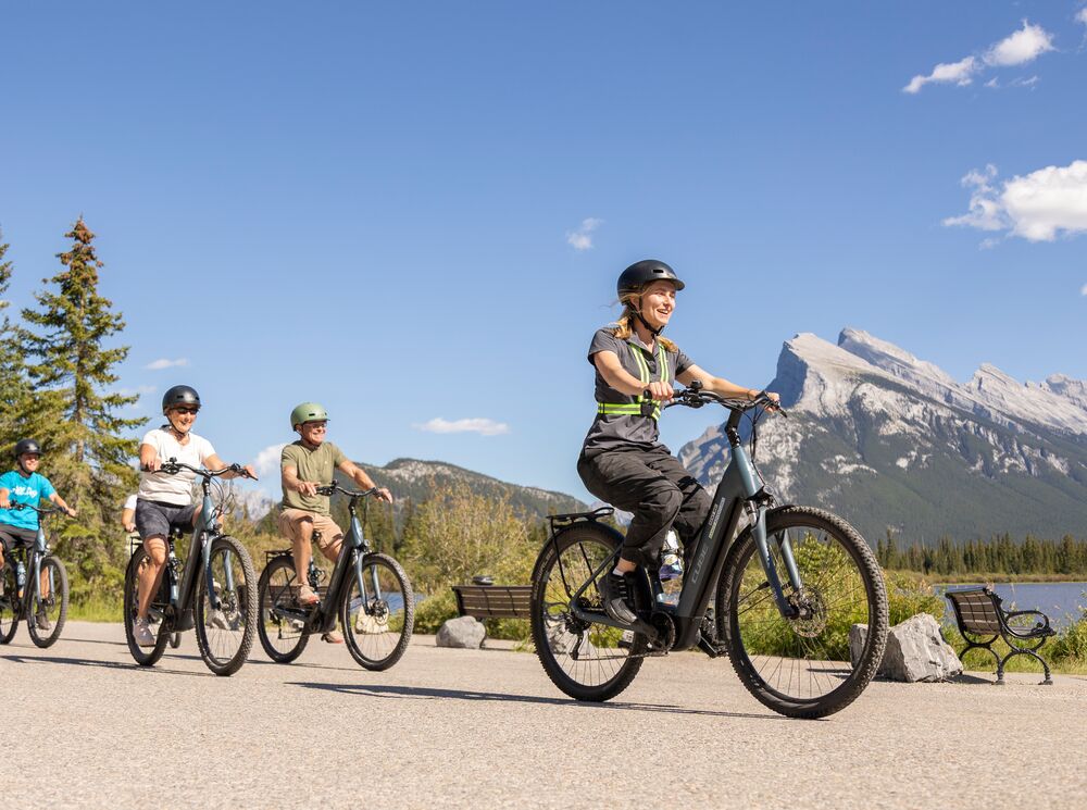 A group of people ride e-bikes in Banff National Park on Vermilion Lakes Road on a bluebird day with Mount Rundle in the background.