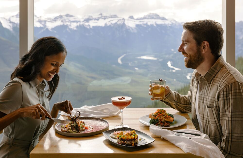 A couple enjoys a meal at the top of the Banff Gondola at Sky Bistro with views of surrounding mountain peaks in Banff National Park