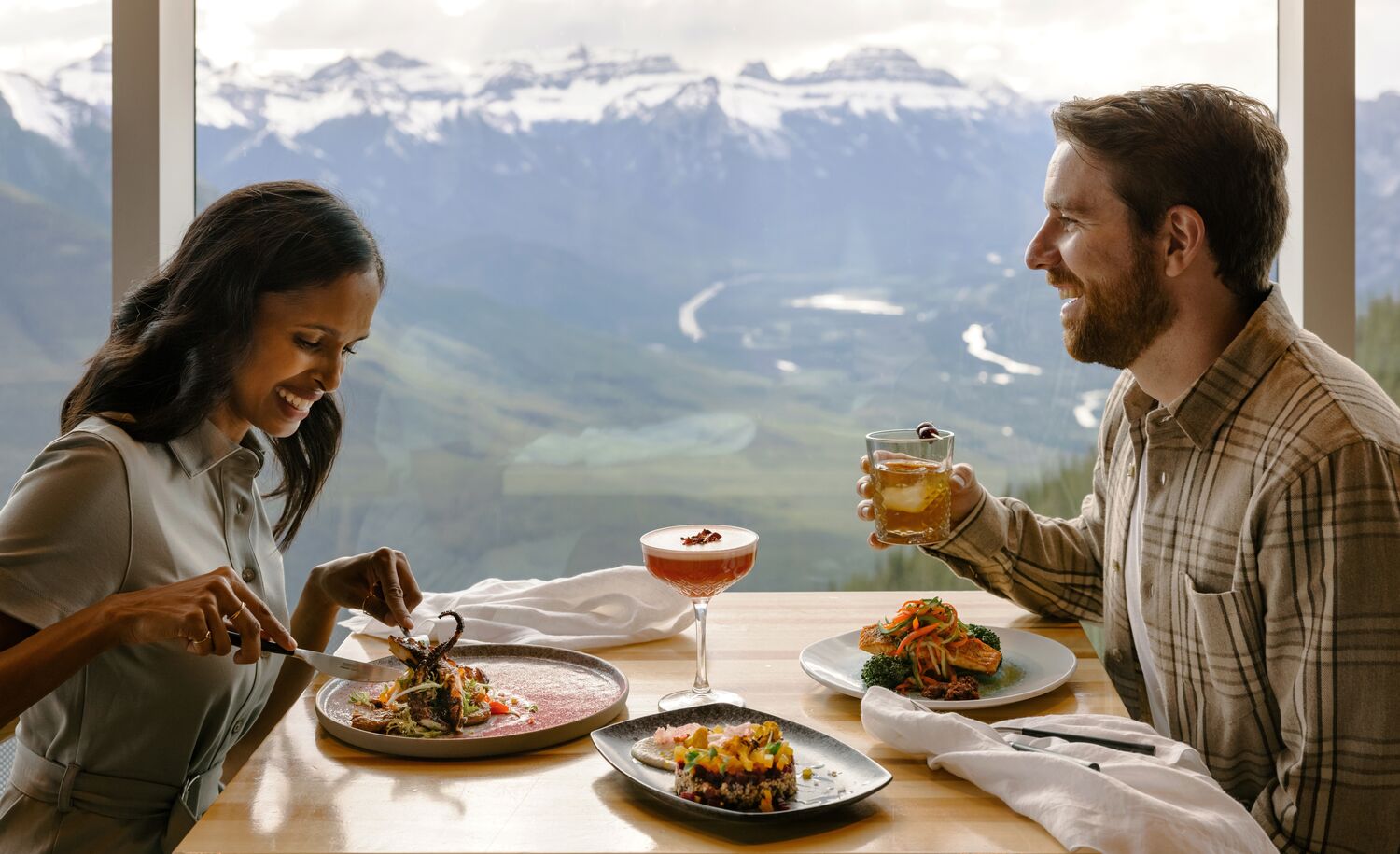 A couple enjoys a meal at the top of the Banff Gondola at Sky Bistro with views of surrounding mountain peaks in Banff National Park