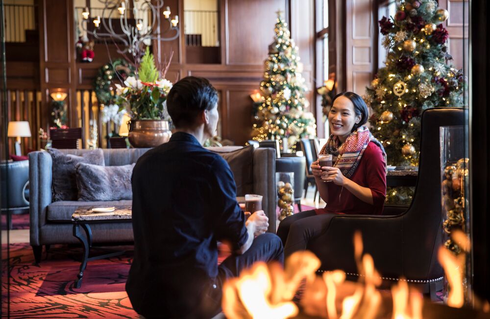 A couple enjoy hot chocolate sitting by a fire place with Christmas trees behind them at the Rimrock Hotel in Banff National Park.