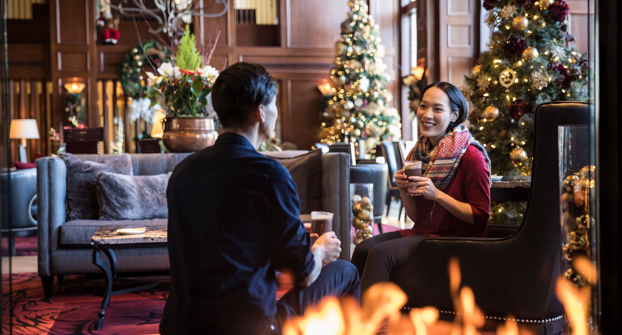 A couple enjoy hot chocolate sitting by a fire place with Christmas trees behind them at the Rimrock Hotel in Banff National Park.