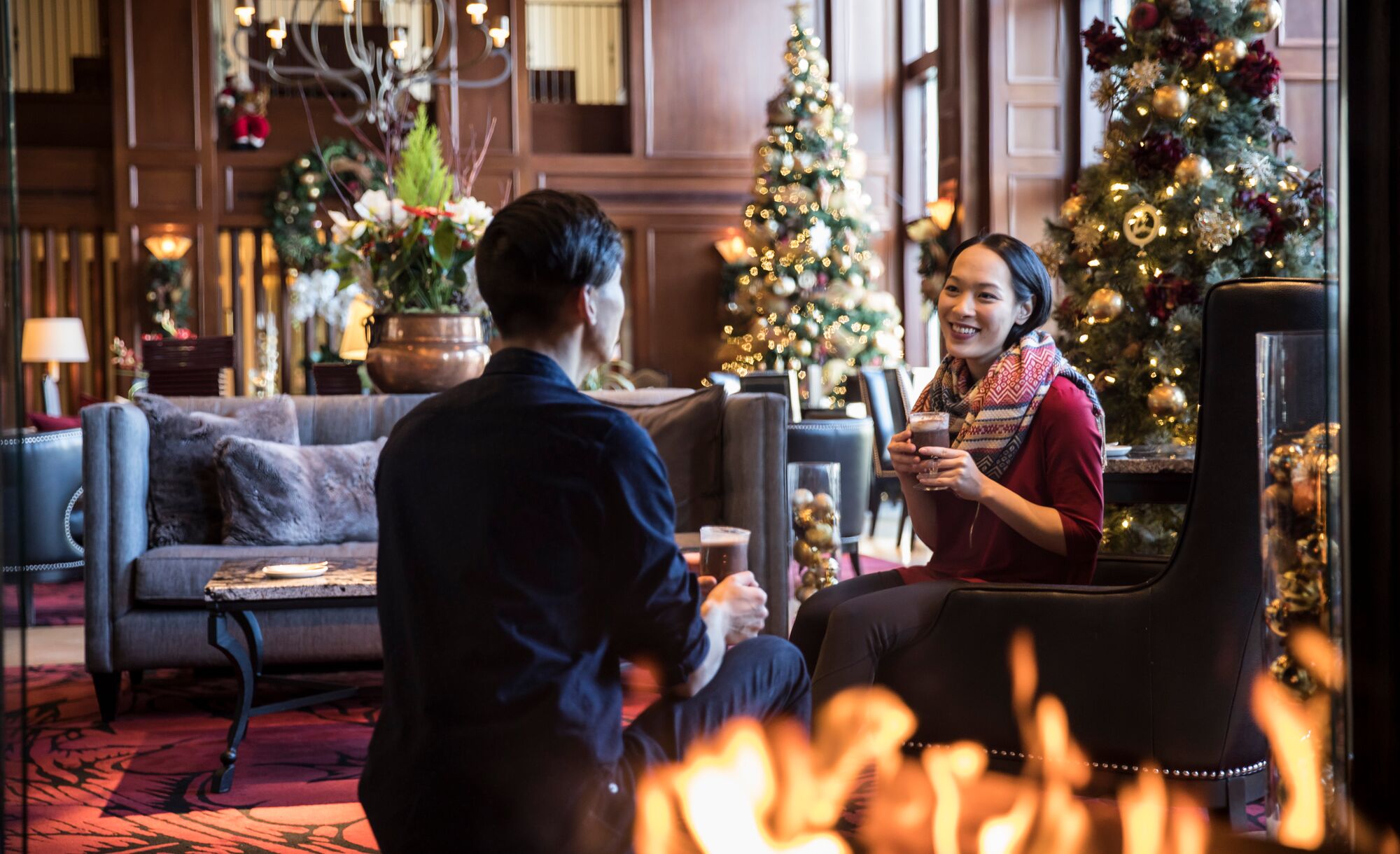 A couple enjoy hot chocolate sitting by a fire place with Christmas trees behind them at the Rimrock Hotel in Banff National Park.