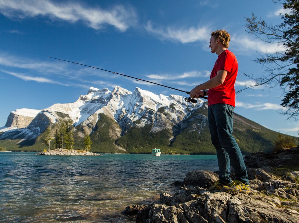 Man fishing from the shore in Lake Minnewanka witha view of the mountains in the background