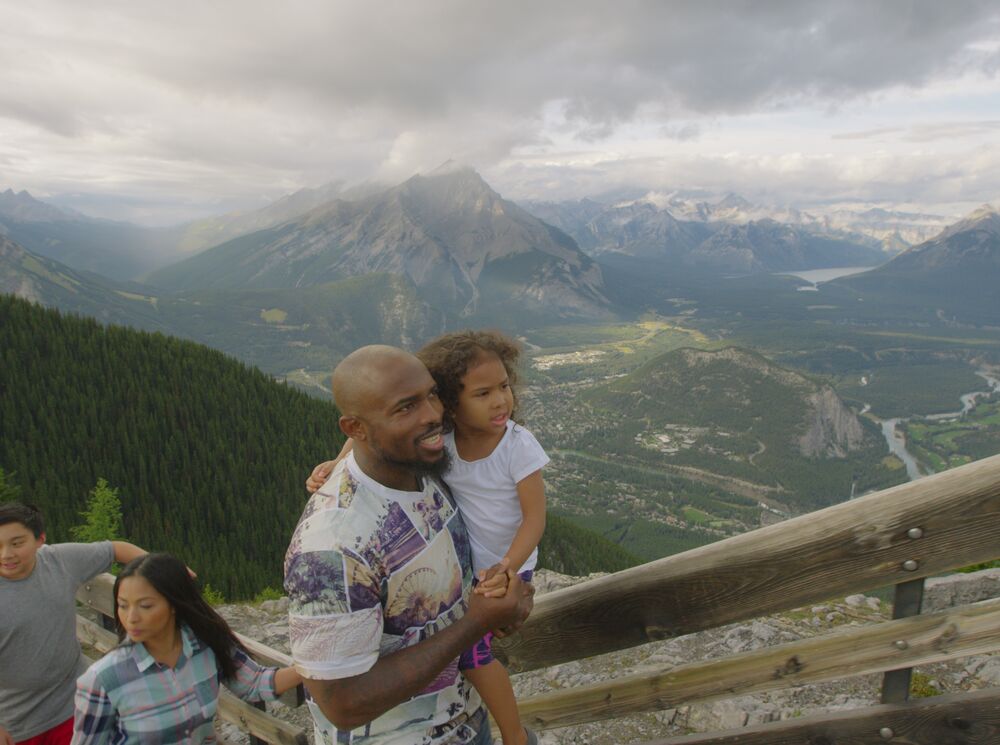 A family walks up the boardwalk at the Banff Gondola with the town of Banff and Cascade Mountain behind them.