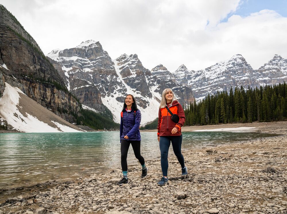 Two friends walk along the lakeshore at Moraine Lake in Banff National Park on a cloudy afternoon with mountains behind them.