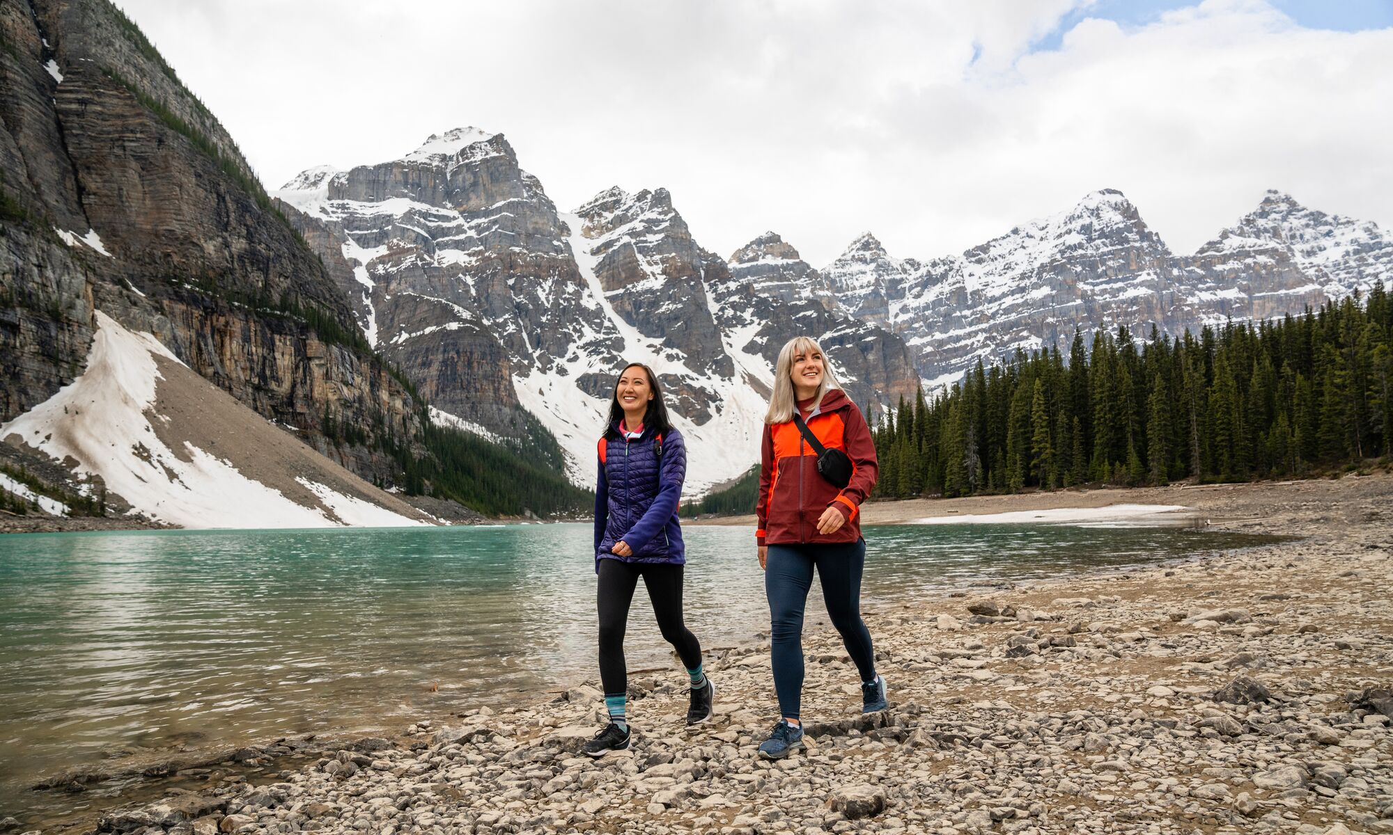 Two friends walk along the lakeshore at Moraine Lake in Banff National Park on a cloudy afternoon with mountains behind them.