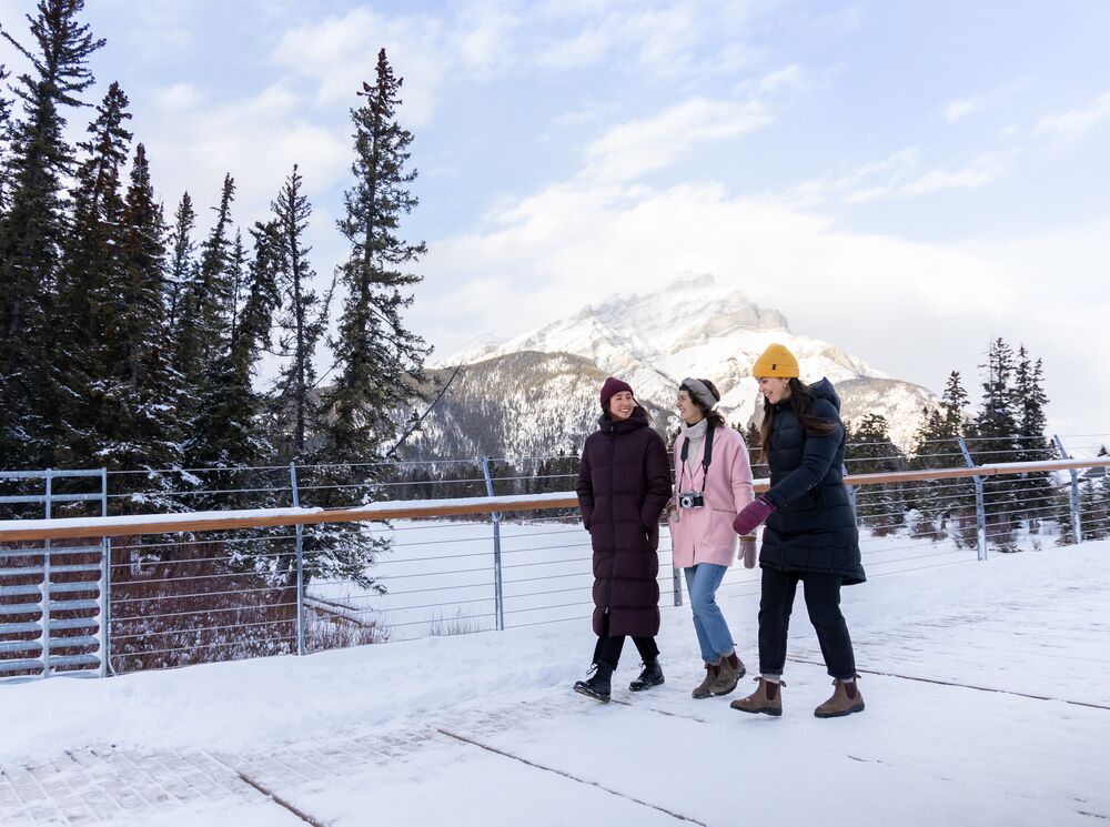 Three friends walk across the Nancy Pauw Bridge in winter with Cascade Mountain behind them and the Bow River below them in Banff National Park.