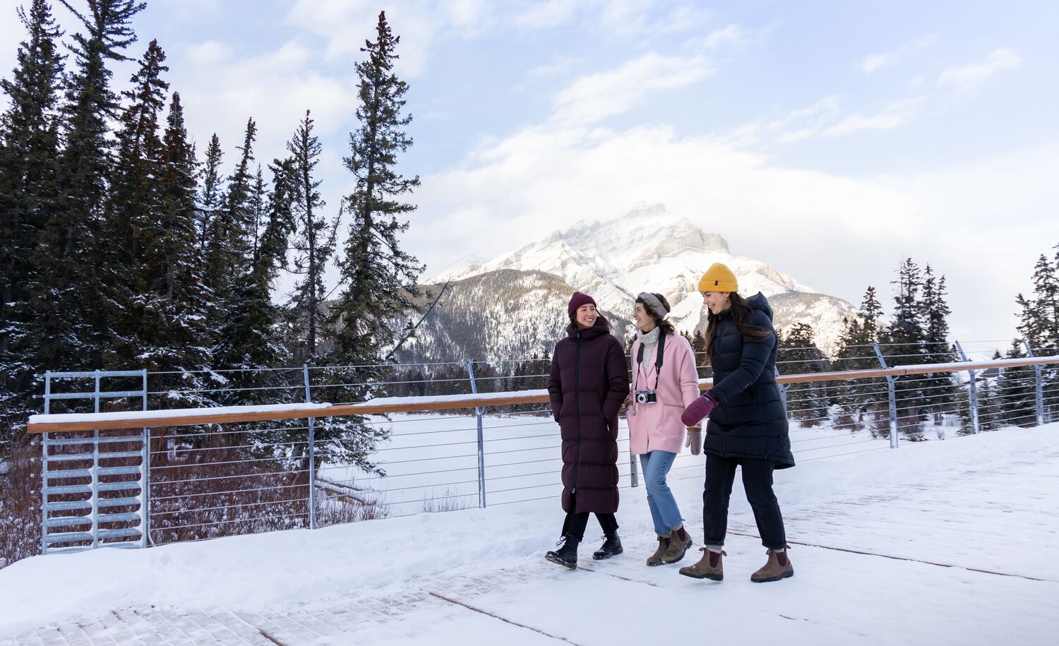 Three friends walk across the Nancy Pauw Bridge in winter with Cascade Mountain behind them and the Bow River below them in Banff National Park.