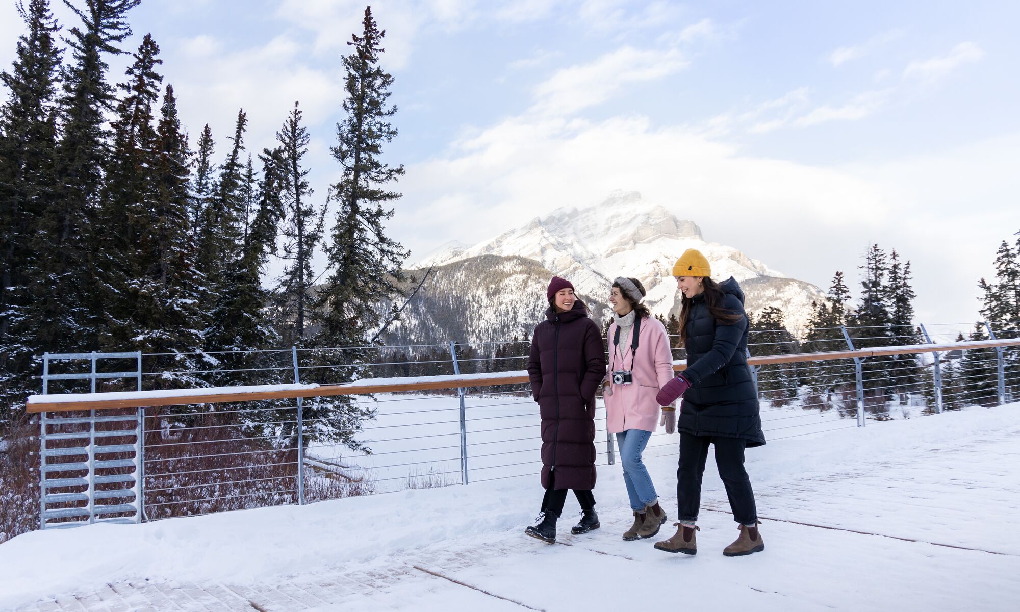 Three friends walk across the Nancy Pauw Bridge in winter with Cascade Mountain behind them and the Bow River below them in Banff National Park.