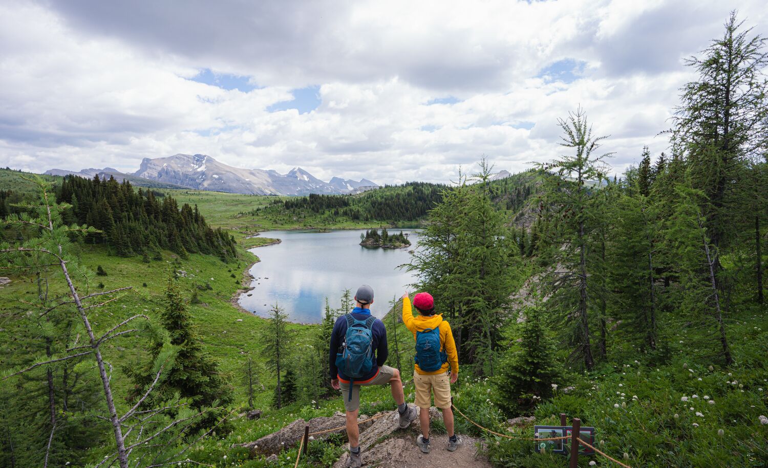 Two hikers look over a lake while on a hiking trail surrounded by trees at Sunshine Meadows near Banff National Park.