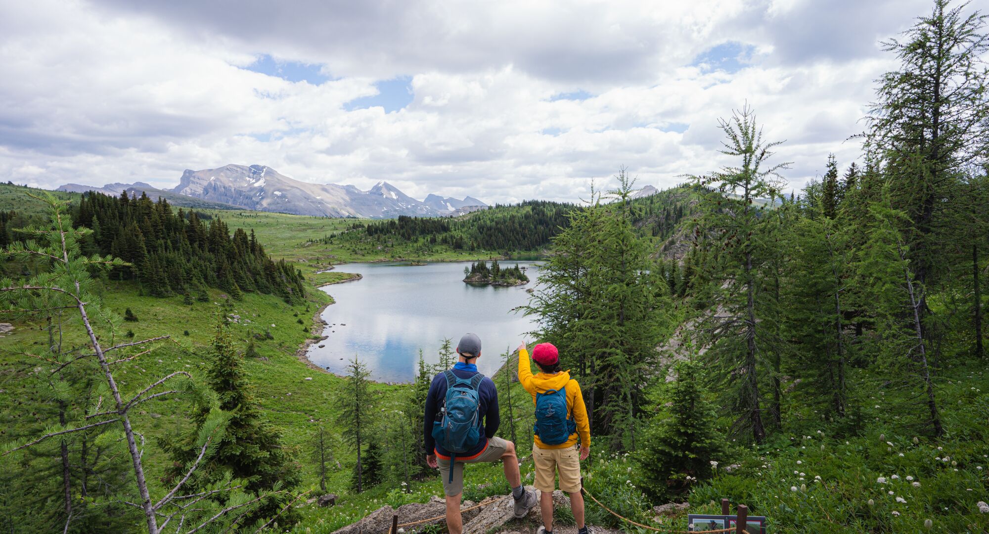 Two hikers look over a lake while on a hiking trail surrounded by trees at Sunshine Meadows near Banff National Park.