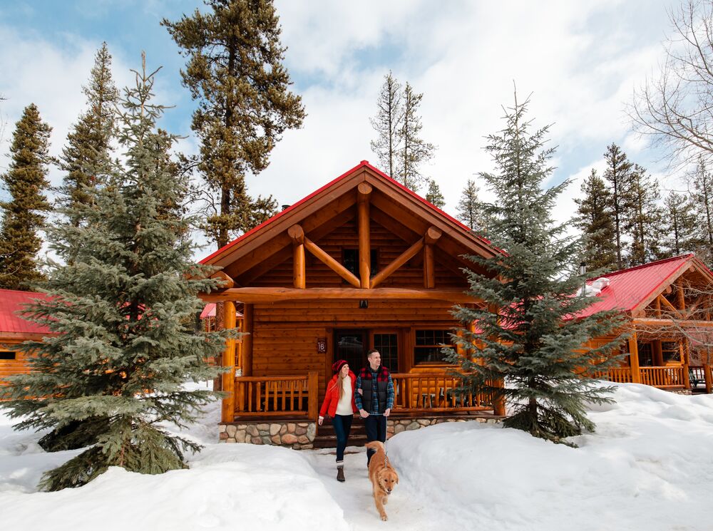 A male and female standing out front of a snowy log cabin with their dog in Banff National Park