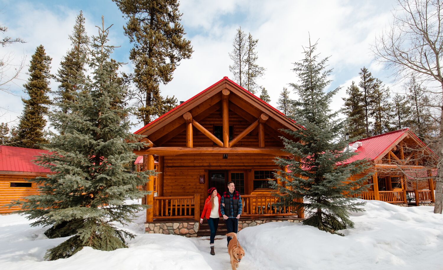 A male and female standing out front of a snowy log cabin with their dog in Banff National Park