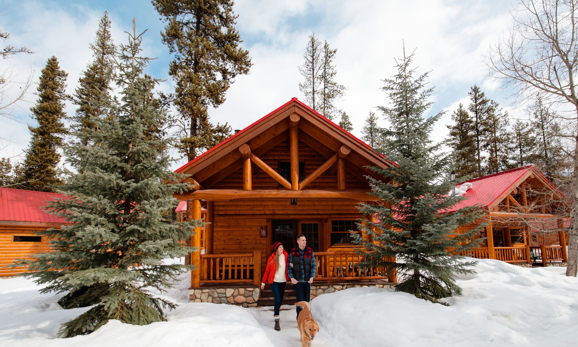 A male and female standing out front of a snowy log cabin with their dog in Banff National Park