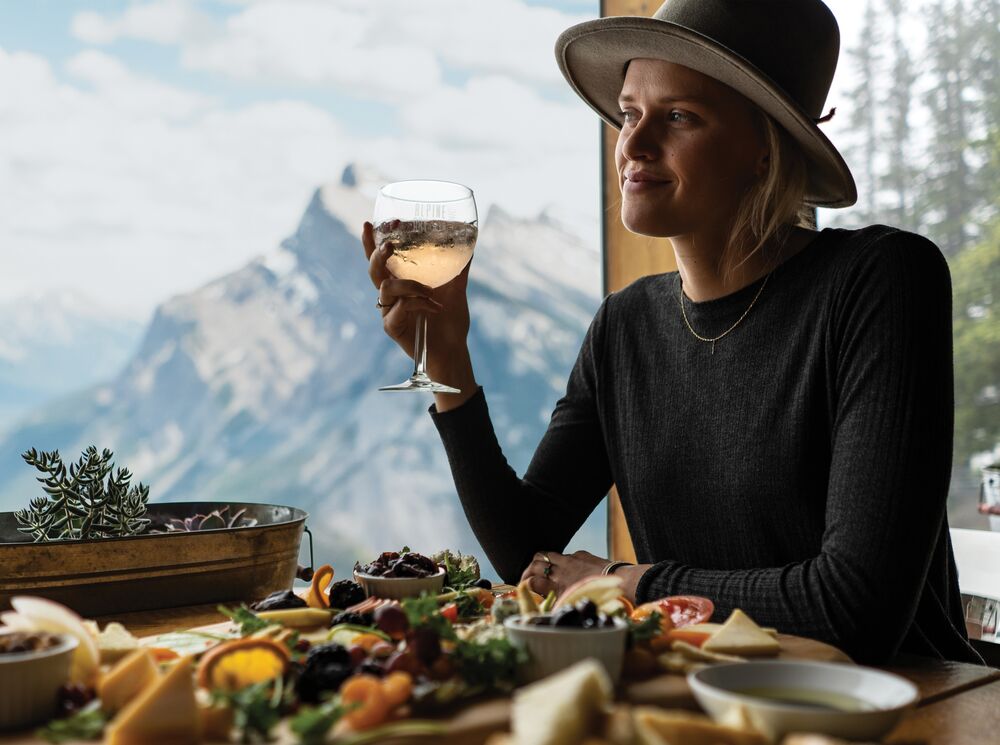 A person in a restaurant with a glass of wine in hand and a charcuterie board with mountain views in the background.