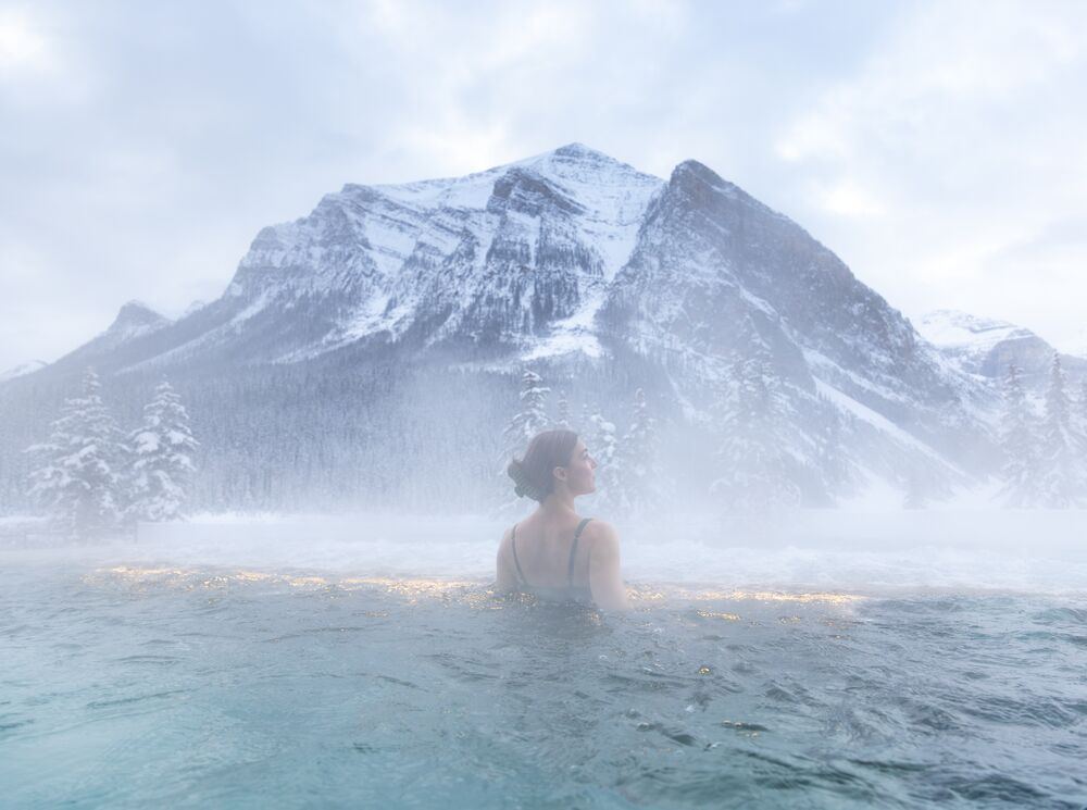 A woman stands alone in an outdoor hot tub with steam around her and a mountain range behind her at the BASIN Spa in Lake Louise in Banff National Park.