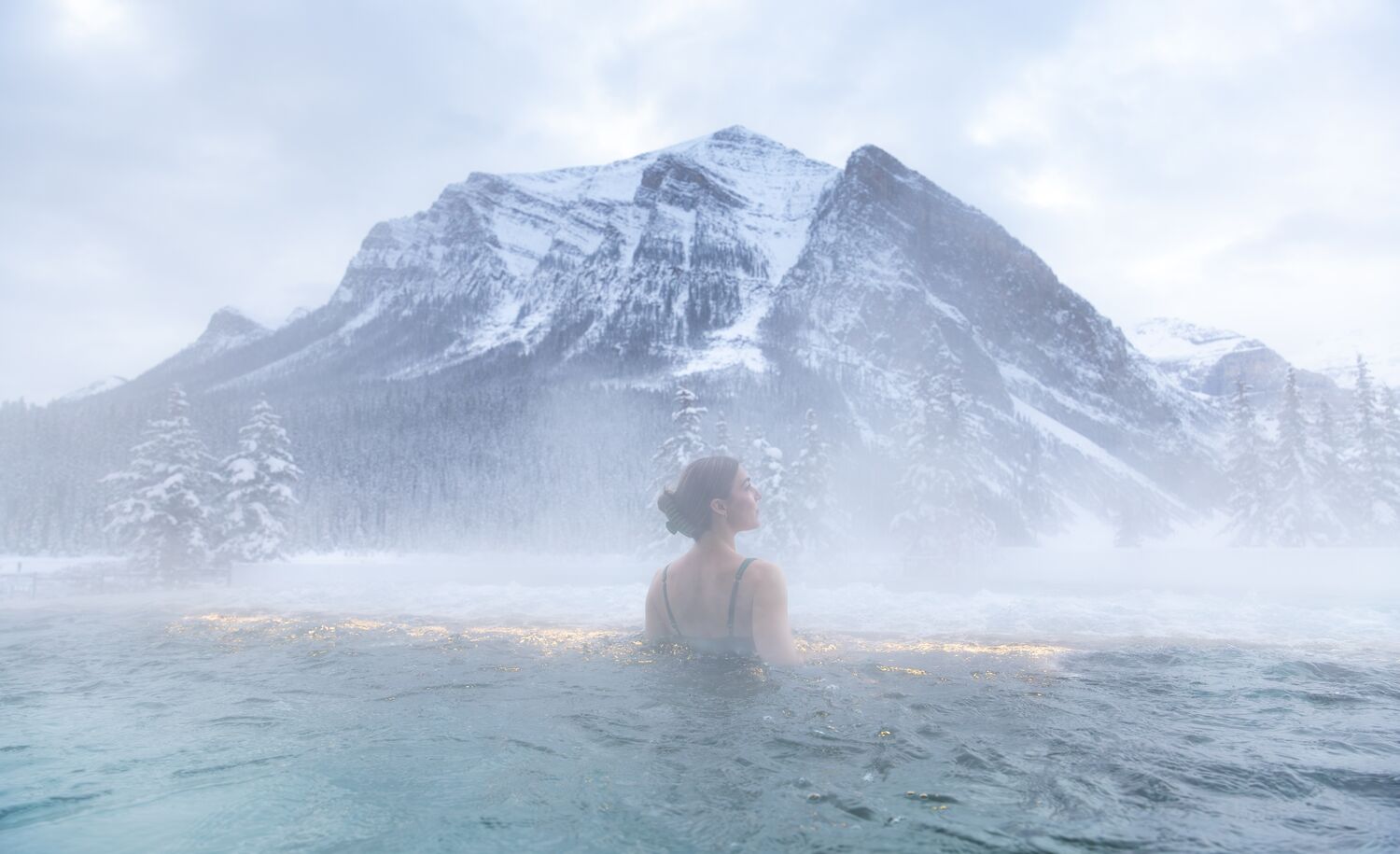 A woman stands alone in an outdoor hot tub with steam around her and a mountain range behind her at the BASIN Spa in Lake Louise in Banff National Park.