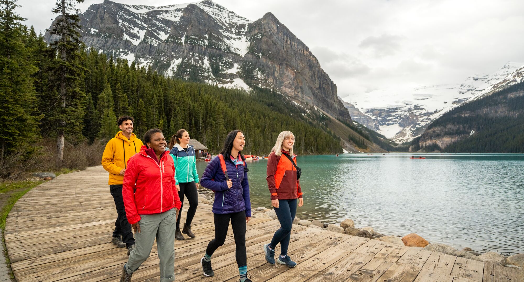 Five friends walk along the boardwalk on the shores of Lake Louise with the lake and mountains behind them in the fall.