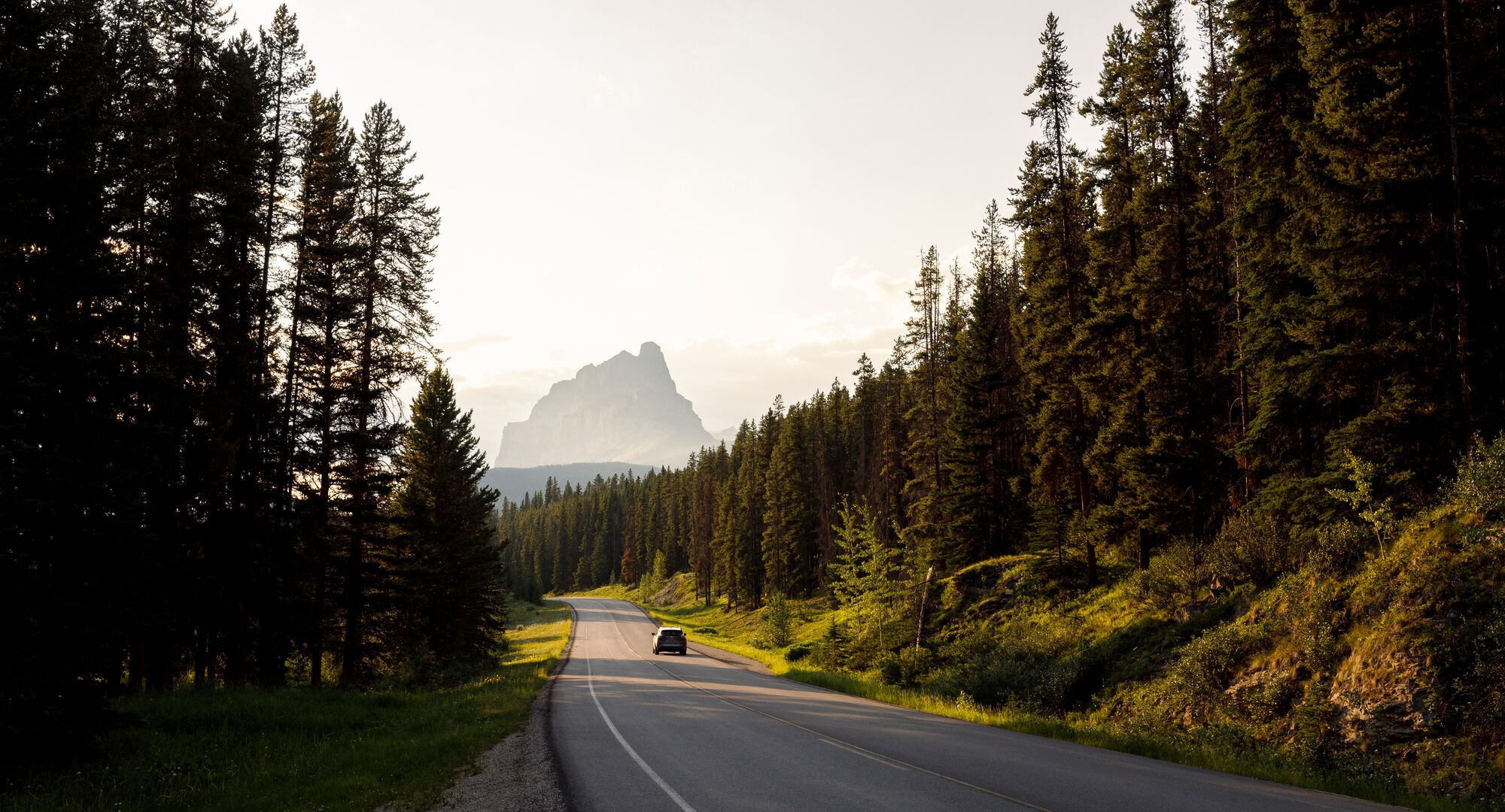 A car driving down a scenic stretch of the Bow Valley Parkway with Castle Mountain in the background