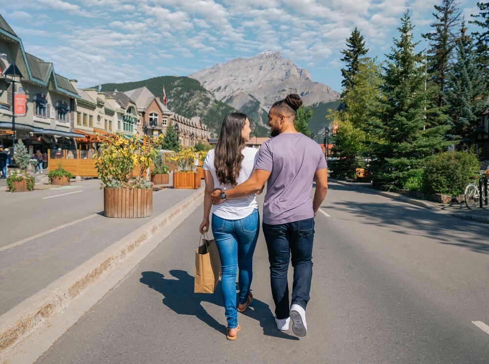 A couple walks in the Banff Avenue Pedestrian Zone in the summer with Cascade Mountain in the background.