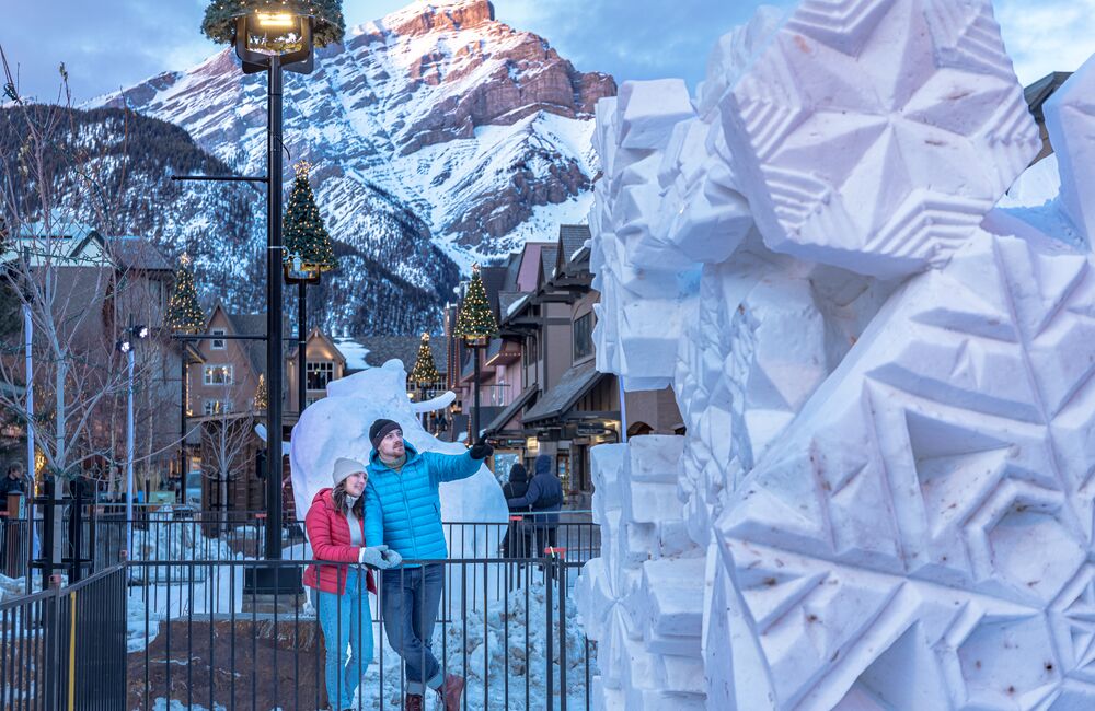 A couple walking in front of a giant snow sculpture with string lights above them in the evening