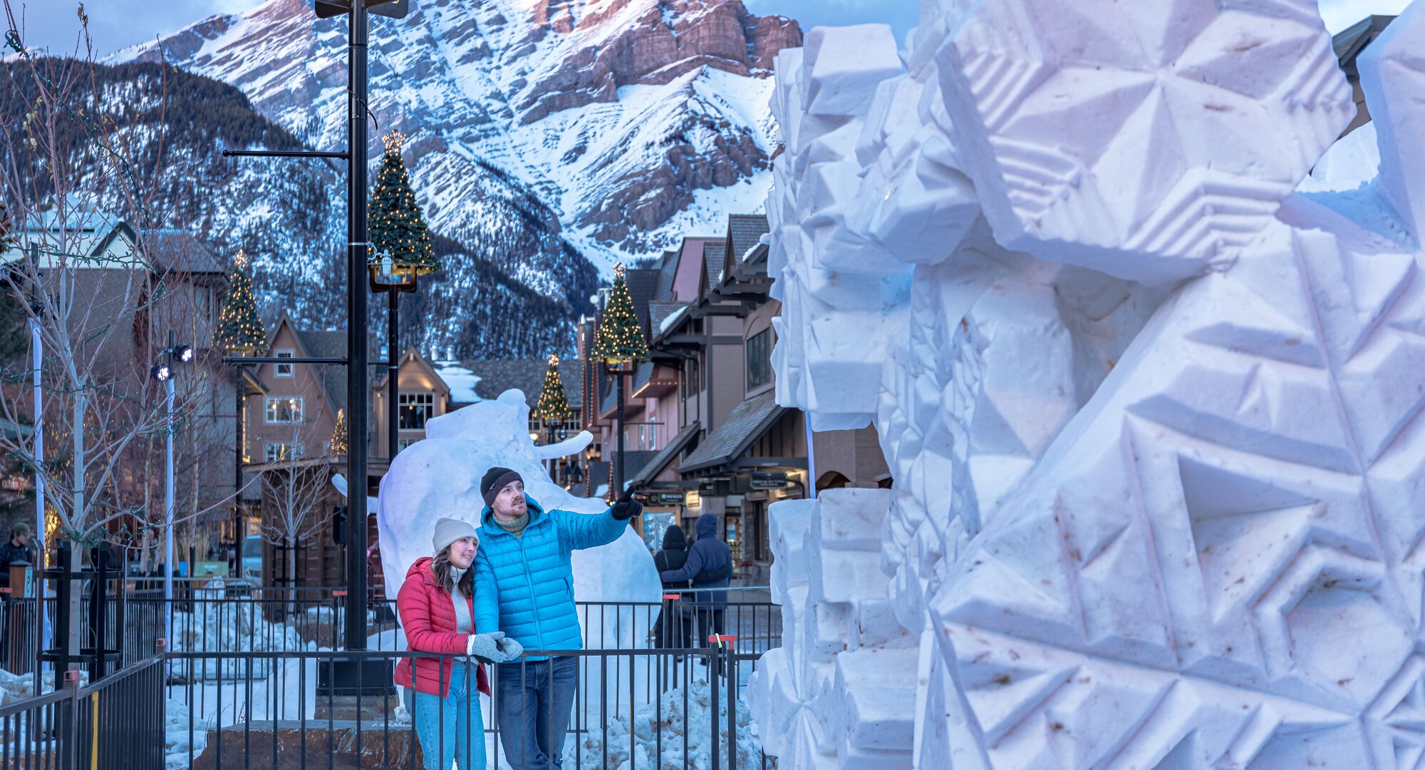 A couple walking in front of a giant snow sculpture with string lights above them in the evening