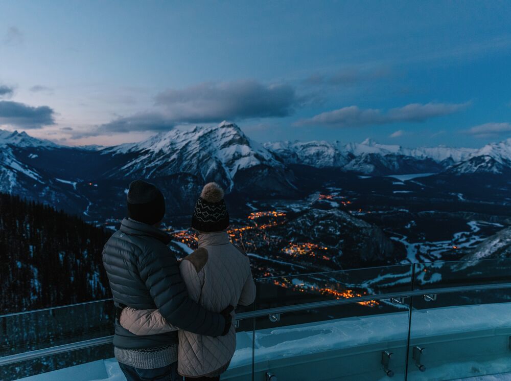 A couple looks out from the observation deck of the Banff Gondola towards Cascade Mountain and the town of Banff in Banff National Park.