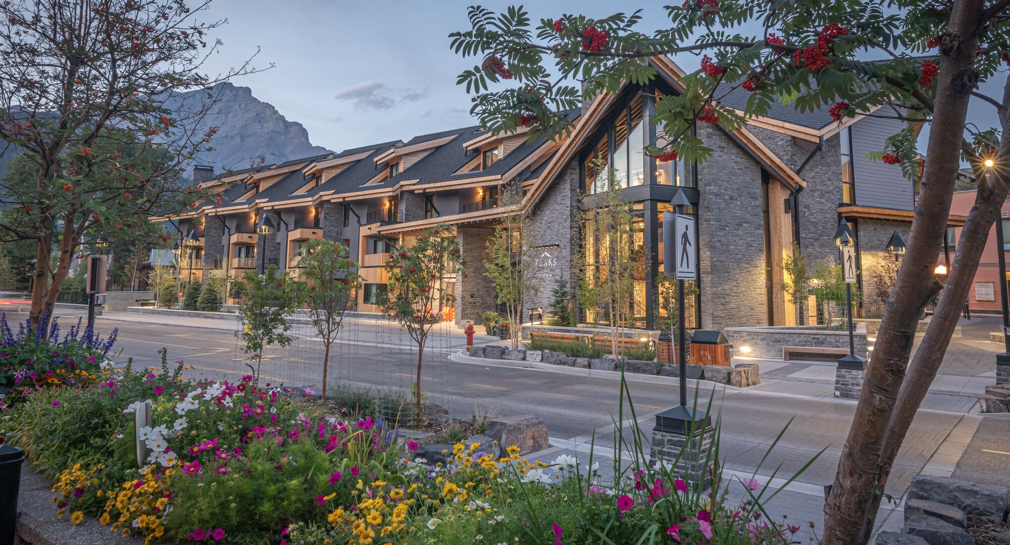 Peaks Hotel & Suites in Banff, featuring contemporary architecture, vibrant flowers, and Mount Rundle in the background at dusk.